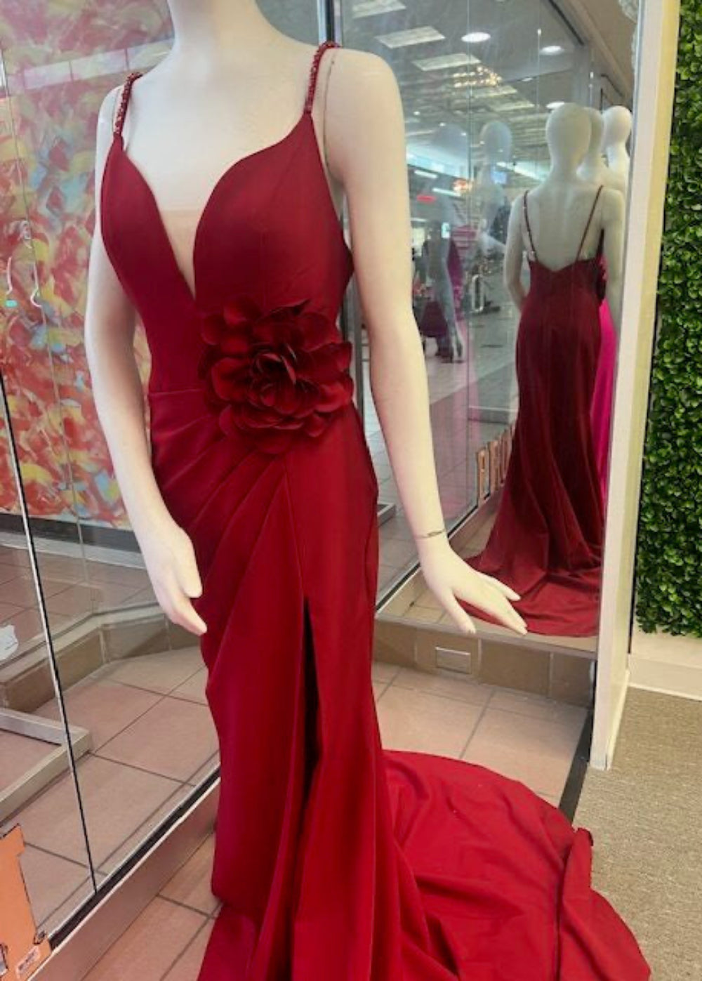 A mannequin wearing a red formal dress with a deep v-neckline and sleeveless design, featuring a floral embellishment at the waist.